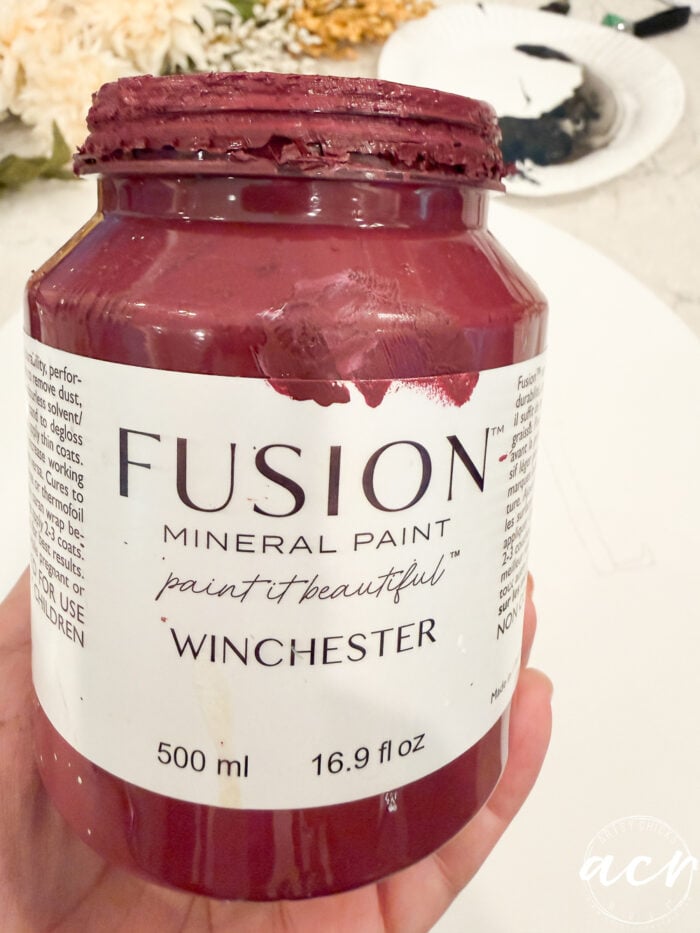 A hand holds an open jar of Fusion Mineral Paint in the shade Winchester, a deep red color perfect for your next Fall Door Hanger. The jar shows some paint around the rim and has a capacity of 500 ml (16.9 fl oz).