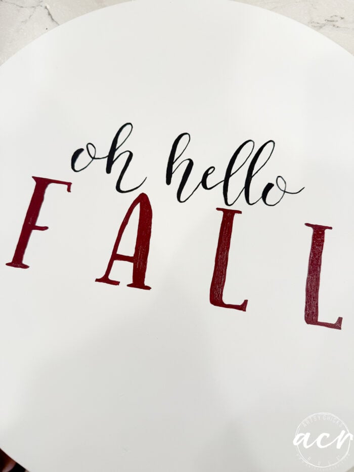 A white circular sign with “oh hello FALL” written on it; “oh hello” is in black script and “FALL” is in large red capital letters. This charming Fall Door Hanger adds a festive touch to your entryway, making it the perfect piece of Fall decor to welcome guests.