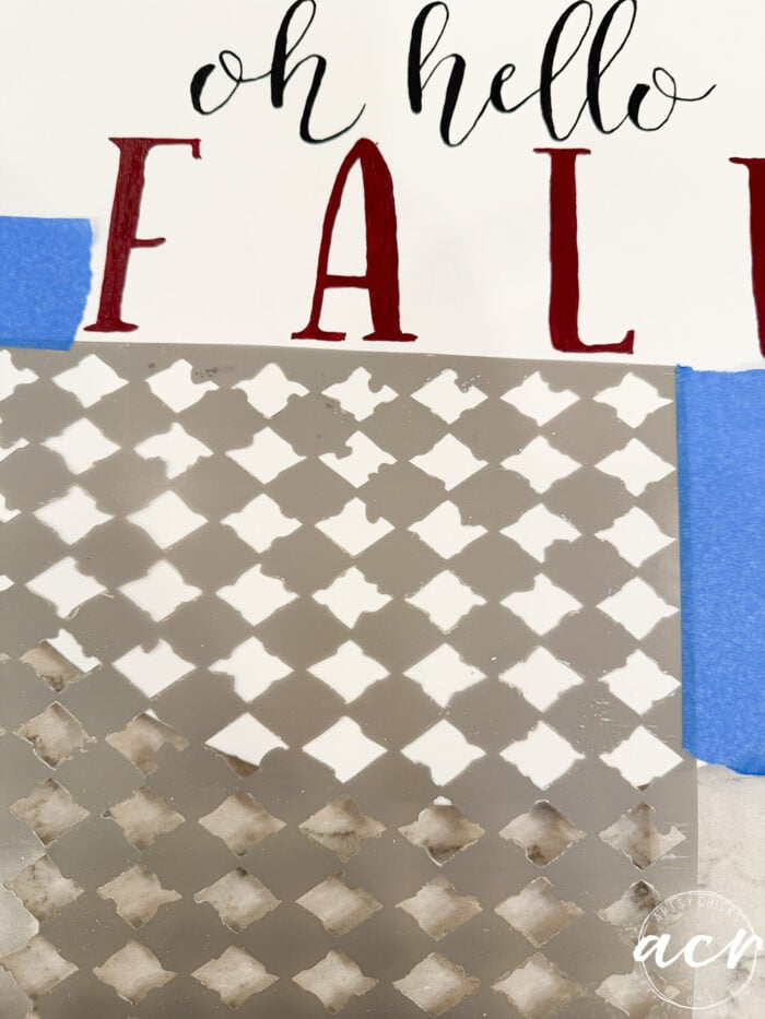 A partially completed fall-themed stenciling project features “oh hello FALL” text above a patterned area secured with blue painter’s tape, perfect for creating a personalized fall door hanger to welcome the season.