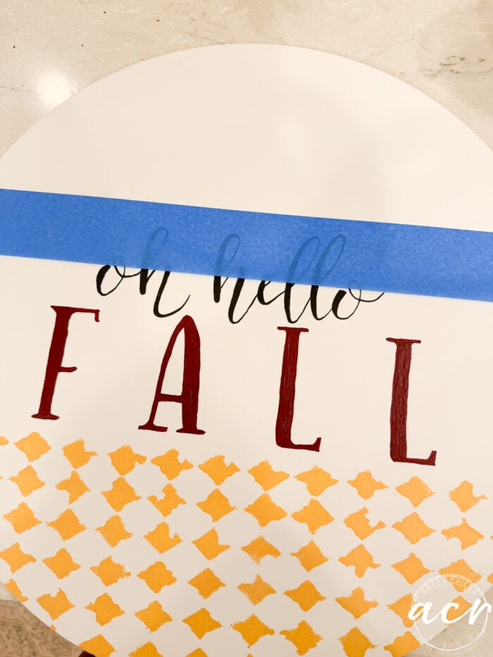 A round Oh Hello Fall Door Hanger sign reads oh hello FALL, with FALL in large red letters. Blue painters tape covers part of the text, and there are yellow diamond patterns at the bottom.