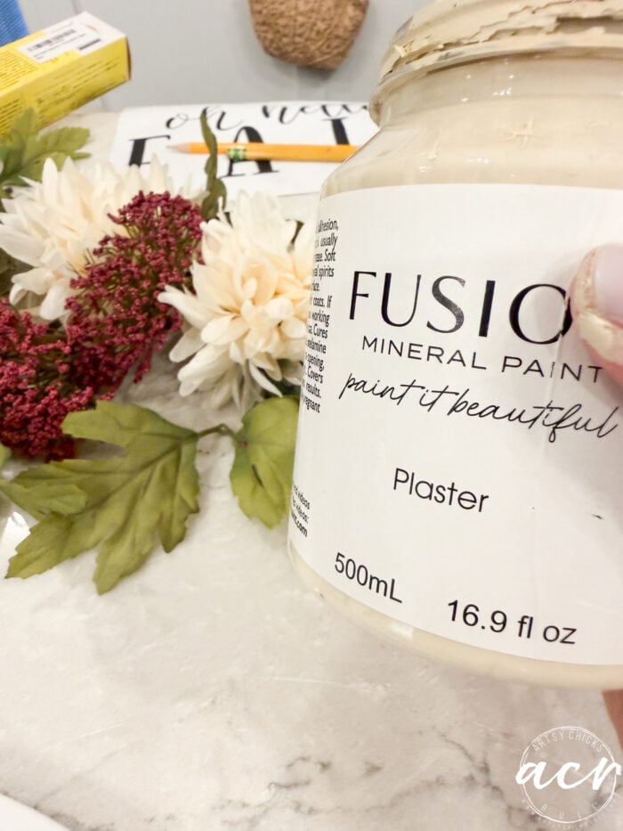 Hand holding a jar of Fusion Mineral Paint in the shade Plaster near artificial flowers and leaves on a marble surface—perfect for your next Oh Hello Fall Door Hanger project. The jar label reads paint it beautiful and displays the size as 500ml (16.9 fl oz).