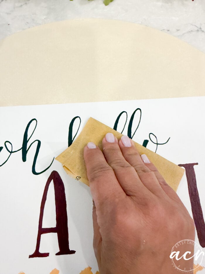 A hand with light pink nail polish holds sandpaper, gently sanding a painted sign with cursive and block letters on a white background, creating the perfect rustic touch for your Hello Fall door decor.