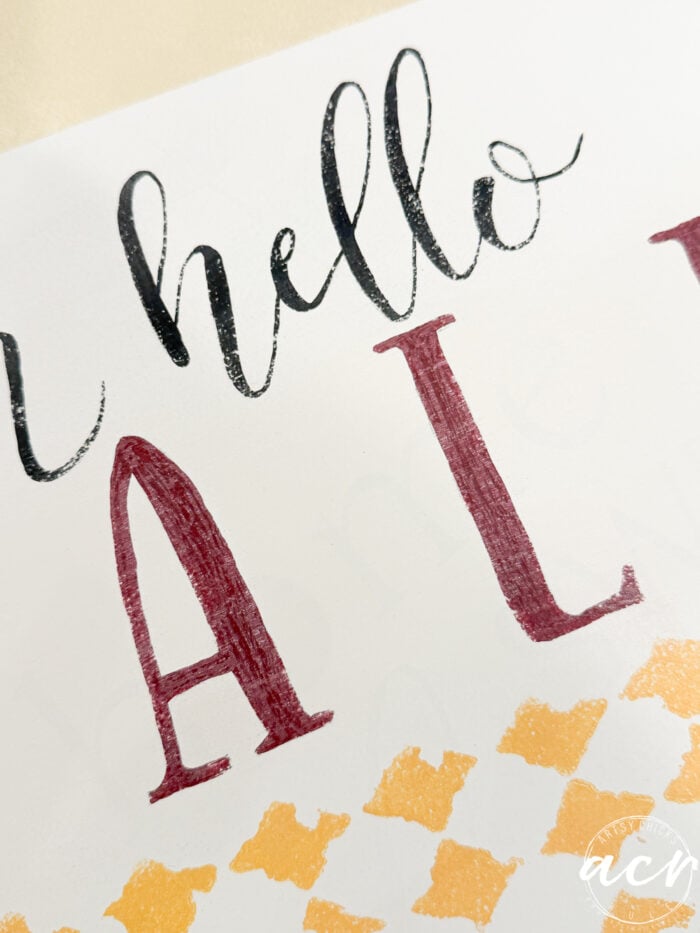 Hand-lettered text on white paper reads Hello Fall in black and maroon, with a gold decorative checkered pattern below—perfect as unique Fall Decor. A white circular logo sits in the bottom right corner.