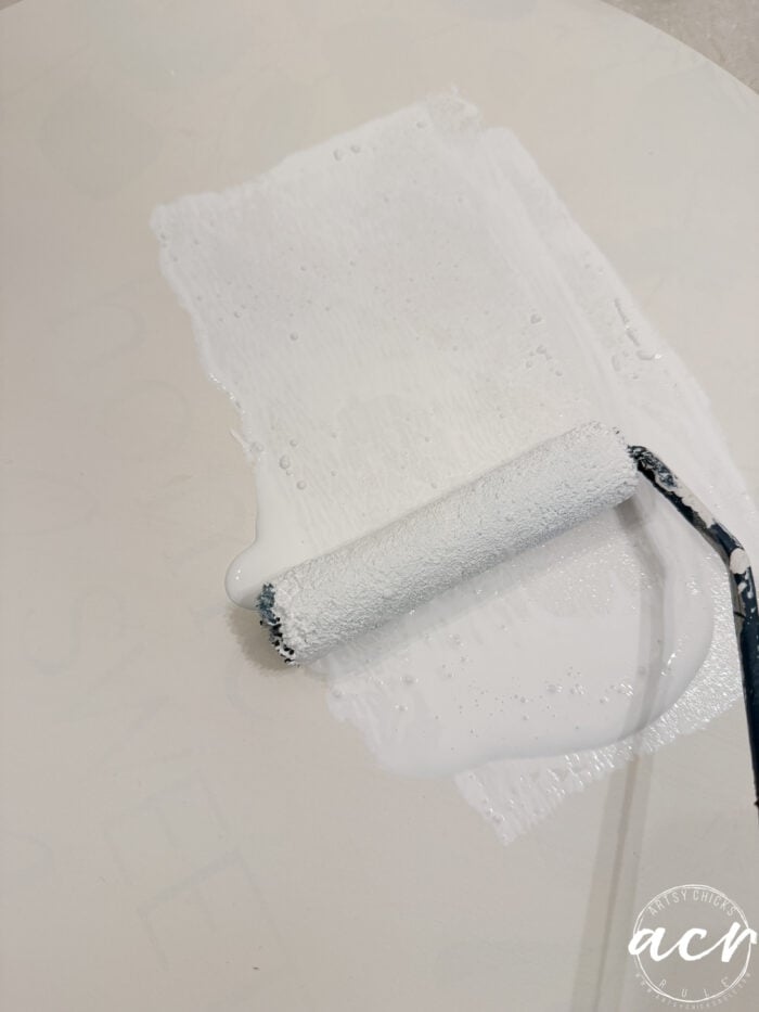 A paint roller coated in white paint is being rolled across a light-colored surface, creating a fresh, even layer of white paint—the perfect base for crafting an inviting Fall Door Hanger.