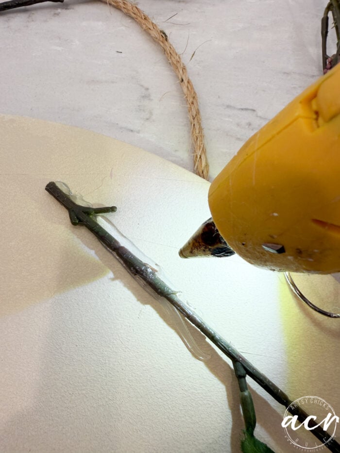 A yellow hot glue gun applies glue to a small branch on a white circular surface, with a piece of twine nearby—perfect for crafting your own fall decor or a charming hello fall door hanger.