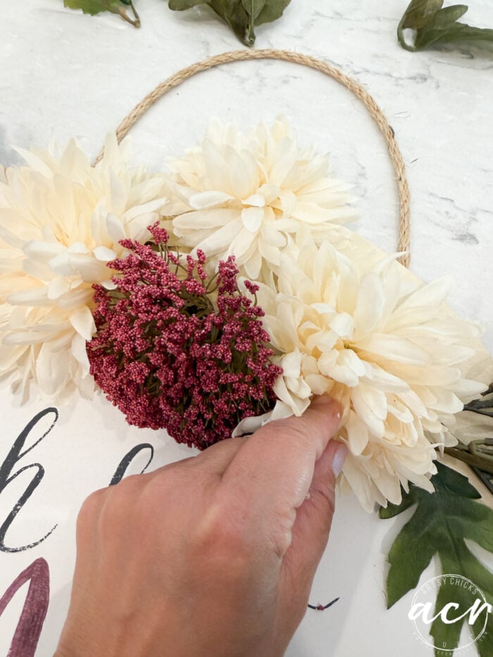 A hand arranges cream and pink artificial flowers on a rope hoop, preparing a beautiful fall door hanger decoration. Green leaves and part of a sign—possibly reading “Oh Hello Fall”—are visible on a white surface.
