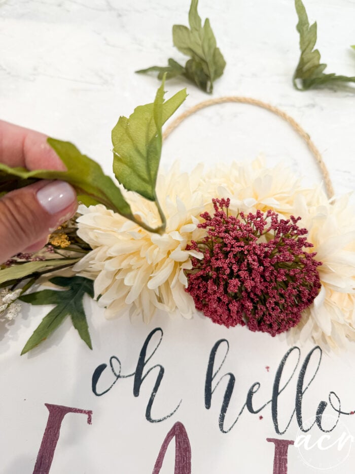 A hand holding a green leaf near a floral wreath with cream and burgundy flowers. The sign below reads “oh hello” in cursive text, making this a charming Fall Door Hanger perfect for welcoming guests with seasonal warmth.