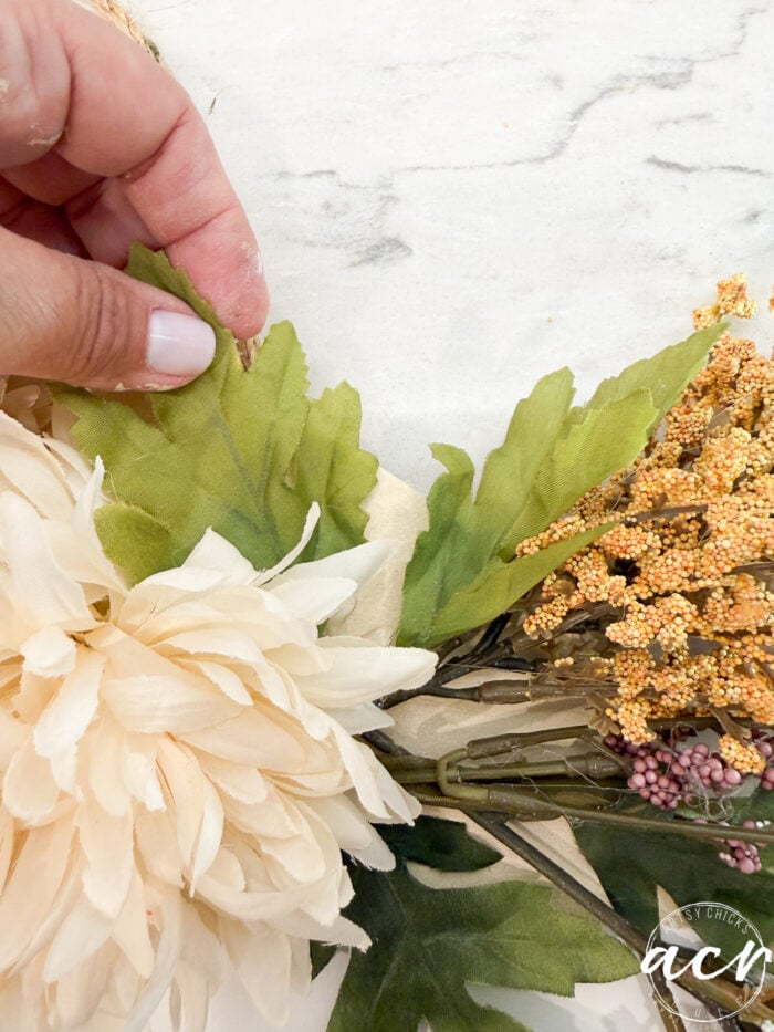 A hand arranges artificial green leaves and beige flowers on a white surface, creating a decorative floral arrangement perfect for a Fall Door Hanger. The background appears to be a marble tabletop.