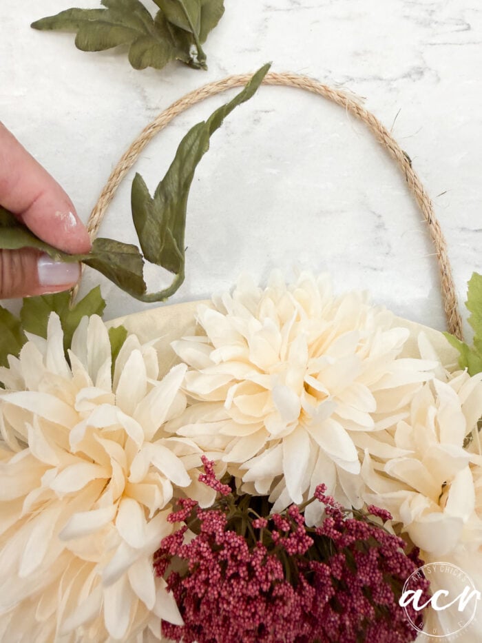 A hand arranges a green leaf among cream and purple artificial flowers attached to a round rope handle on a white surface, creating a charming fall door hanger.