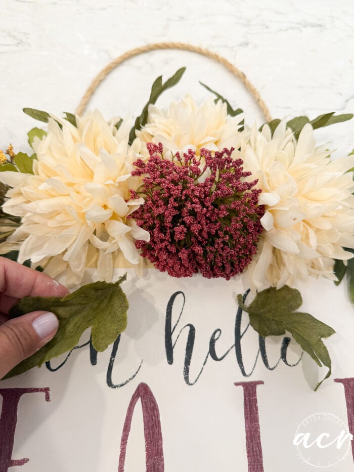 A hand arranges green leaves on a floral wreath with cream and burgundy flowers above a sign that says Oh Hello Fall in cursive letters, creating the perfect Fall Door Hanger to welcome guests.