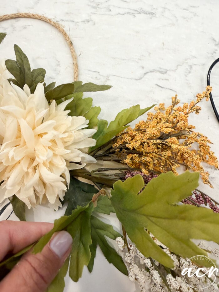 A hand holding green artificial leaves, arranging them with a cream flower and golden accents on a marble surface—a rope or wreath base is partially visible, creating the perfect foundation for a charming Fall Door Hanger.