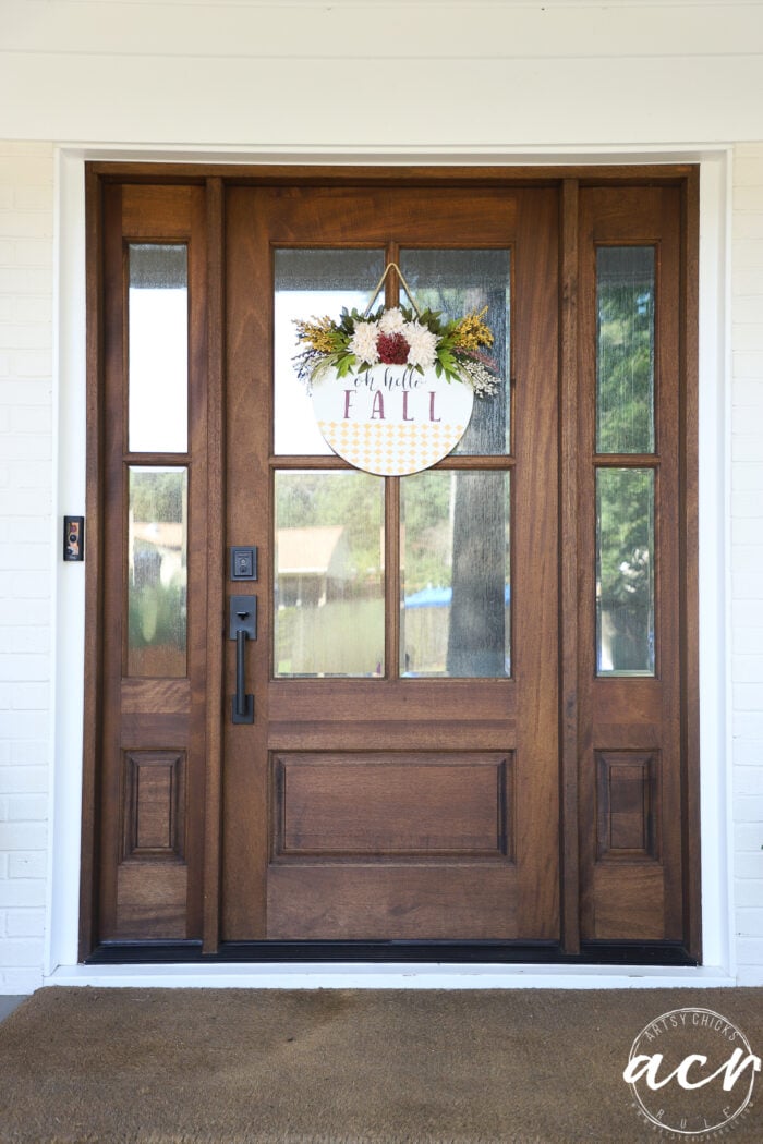 A wooden front door with glass panels and a round Fall Door Hanger that says “Hey there FALL” hanging at the center, decorated with autumn leaves and flowers.