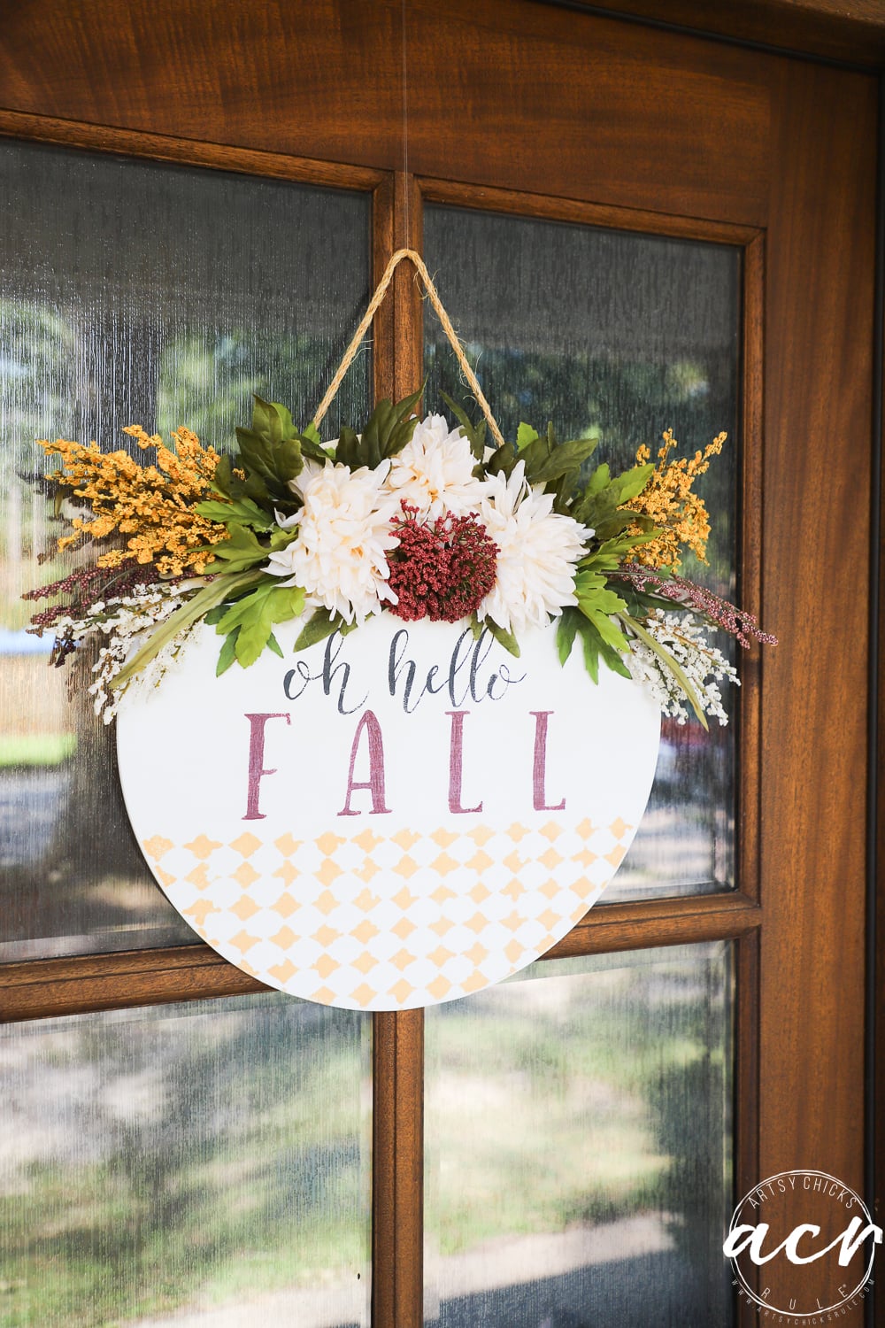 A round wooden sign, perfect as a Fall Door Hanger, features elegant flowers and leaves surrounding the words “oh hello FALL” in flowing script. Hanging on a door, this charming piece is accented by a gold diamond pattern along the bottom, creating a warm and inviting seasonal welcome.