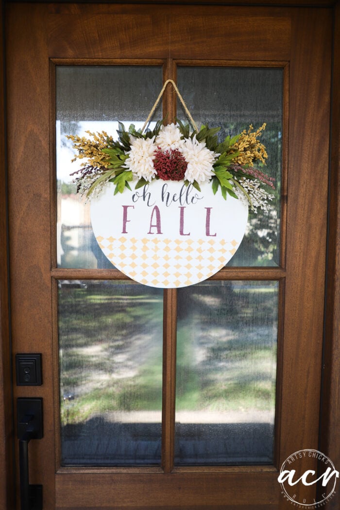 A wooden door with a glass panel displays an Oh Hello Fall Door Hanger—a round sign reading “oh hello FALL,” decorated with white and burgundy flowers and greenery, hanging from a rope.