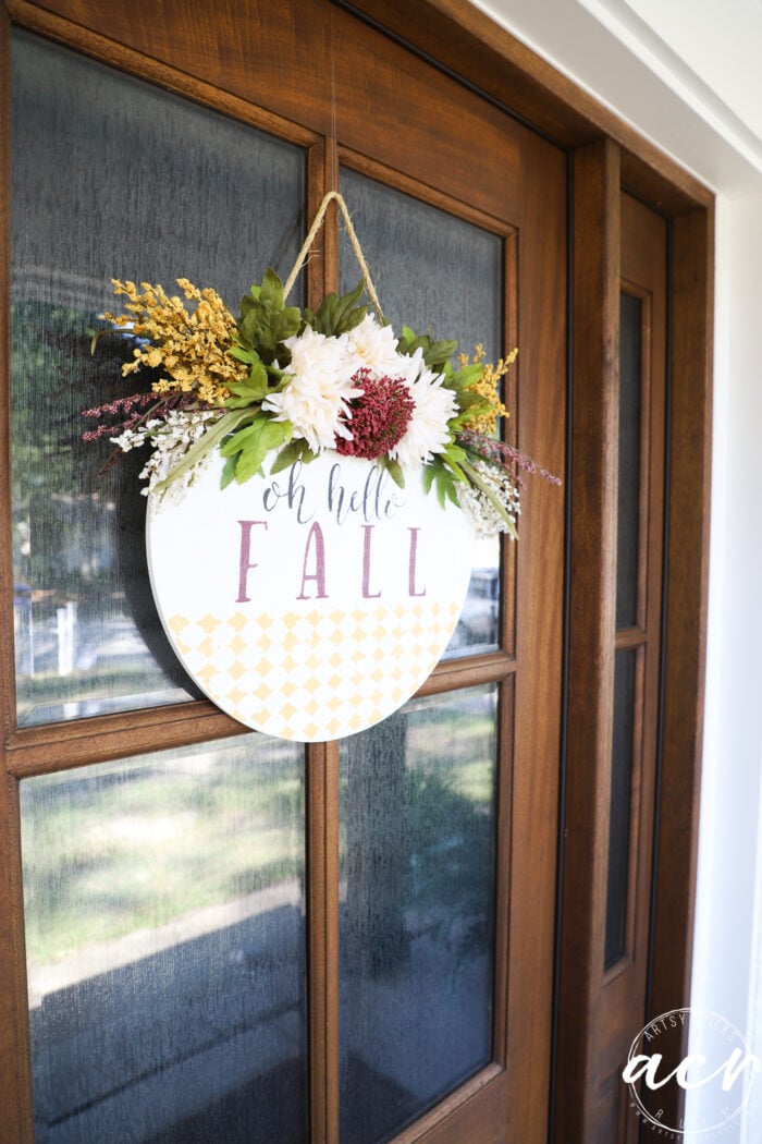 A round sign with flowers and greenery hangs on a wooden door, reading oh hello FALL in decorative letters—a charming Fall Door Hanger that adds the perfect touch of seasonal door decor.