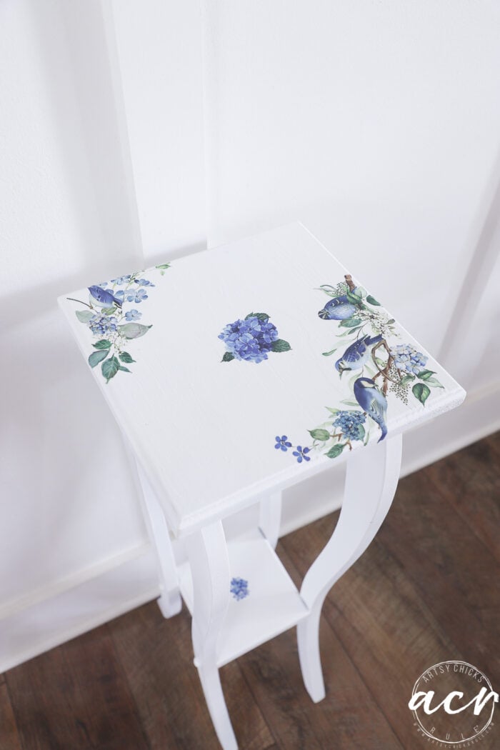 A petite table, this small white side table features blue and purple floral and bird designs on the tabletop, placed gracefully on a wooden floor against a white wall—an eye-catching result of a charming furniture makeover.