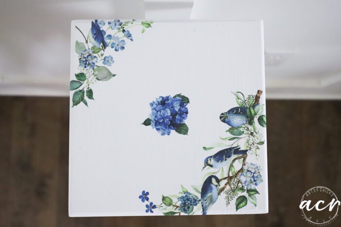 A white tabletop, perfect for a petite table makeover, is decorated with blue flowers and birds in two corners, viewed from above. There is a circular acn watermark in the bottom right corner. The background floor is dark brown—an inviting touch for any furniture restoration project.