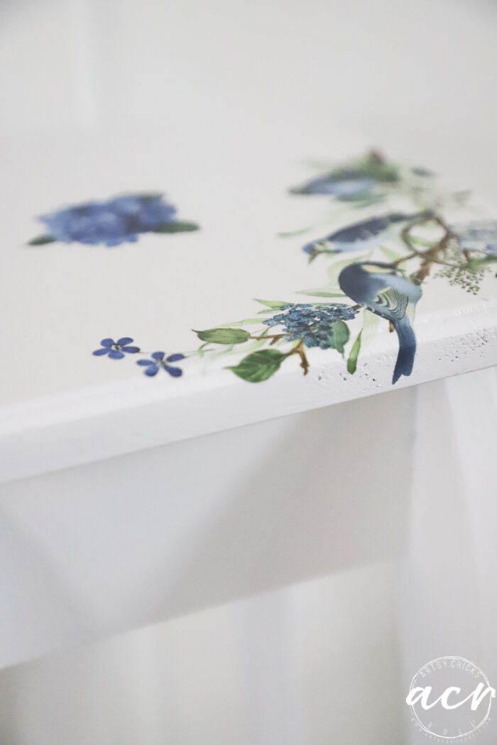 A white petite table corner decorated with a painted blue bird, blue flowers, and green leaves. The design is delicate and detailed, adding a touch of nature to this charming furniture makeover.