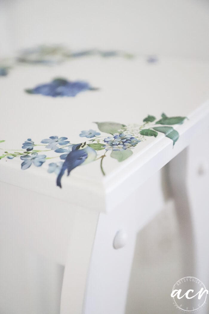 A white petite table with blue and green floral designs on the corner of the surface. The flowers appear delicate and detailed, adding a decorative touch to the minimalist furniture—perfect for anyone seeking a charming furniture makeover.