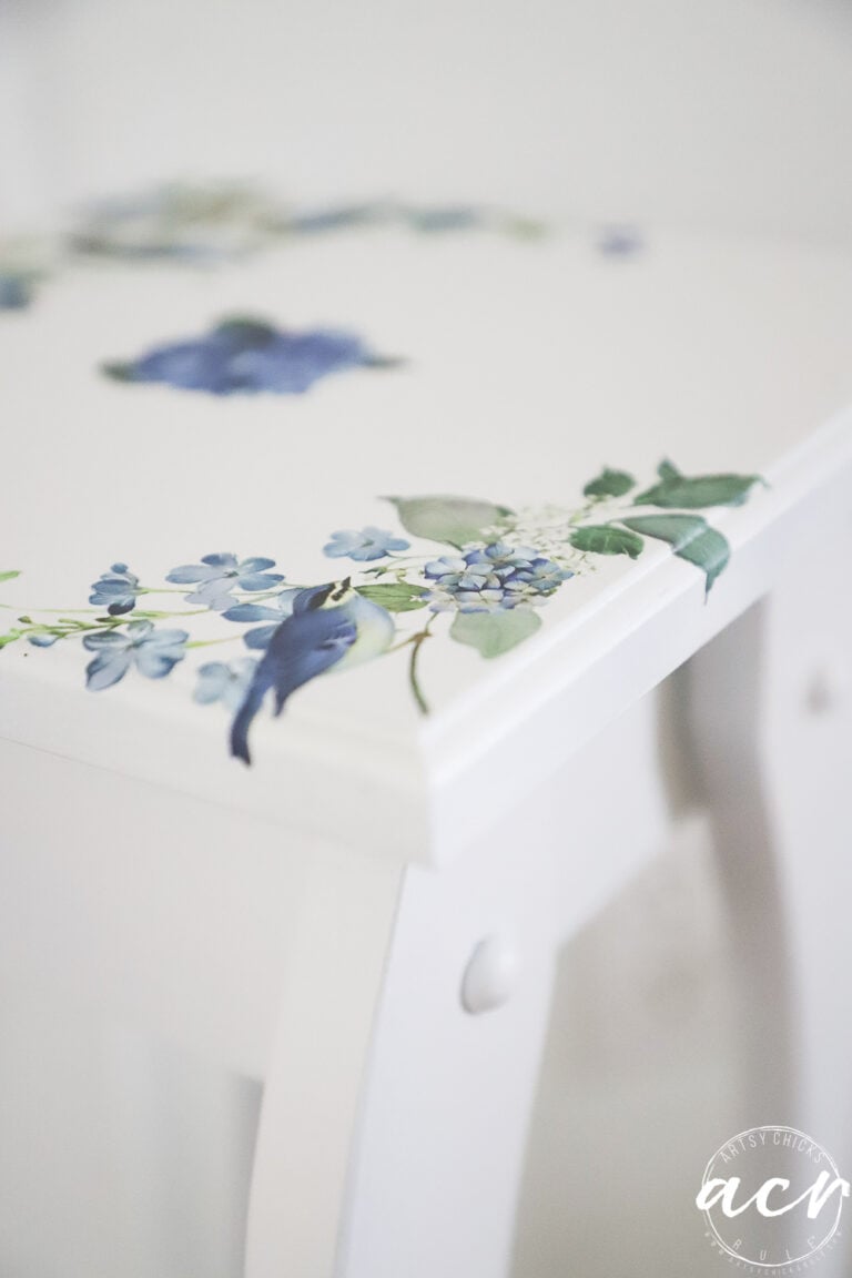 A white petite table with blue and green floral designs on the corner of the surface. The flowers appear delicate and detailed, adding a decorative touch to the minimalist furniture—perfect for anyone seeking a charming furniture makeover.