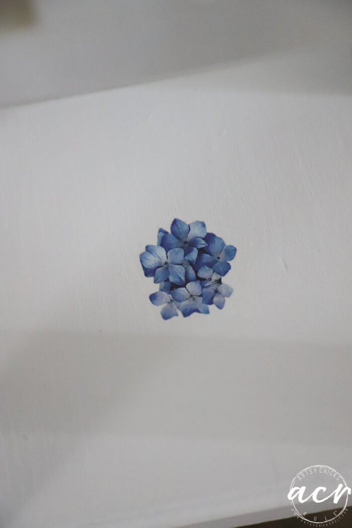 A small cluster of blue hydrangea petals is painted on a white surface, reminiscent of a petite table makeover, with the artist’s watermark in the lower right corner.