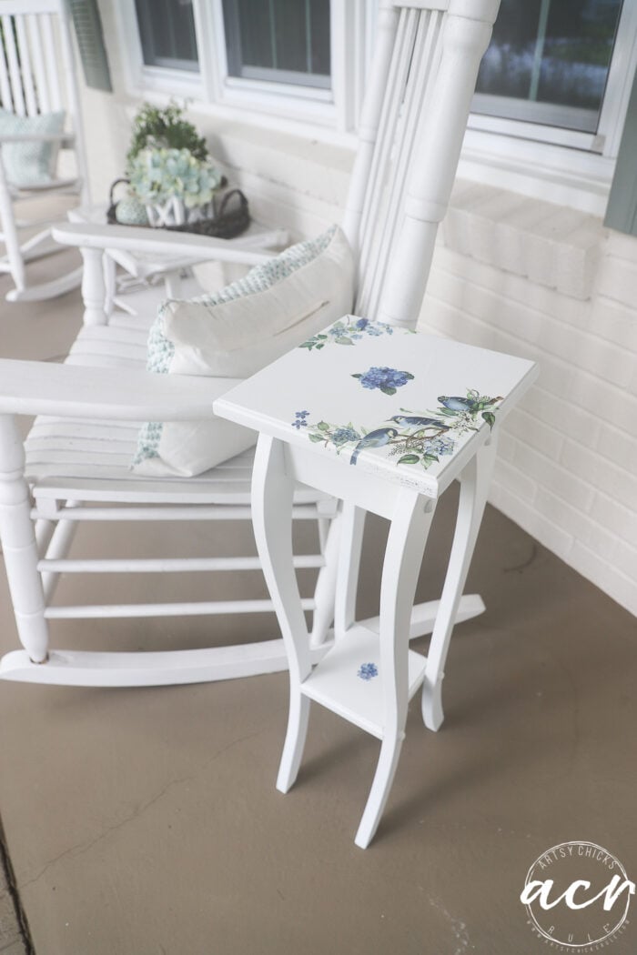 This petite table with curved legs sits elegantly on a porch, showcasing a delightful furniture makeover with blue floral and bird designs. Next to it, a white rocking chair with a pillow adds charm, while a plant arrangement completes the inviting scene.