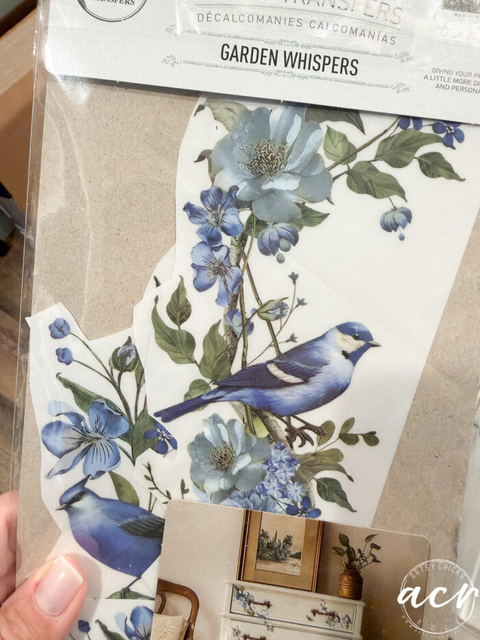 A hand holds a package of floral and bluebird wall decals labeled Garden Whispers; an example of the decals used decoratively is shown at the bottom right corner of the packaging, perfect for adding a charming touch to your next furniture makeover or petite table project.
