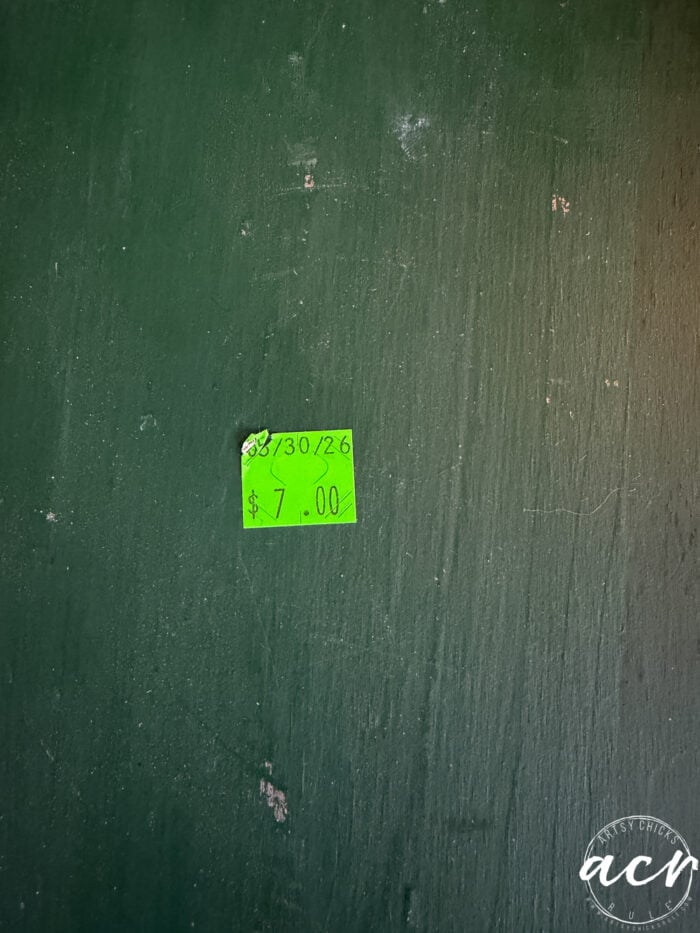 A bright green price tag on a dark green wall shows the date 03/30/26 and a price of $7.00—perhaps signaling the start of a petite table makeover. The wall has visible texture and a watermark with the initials acr in the bottom right corner.