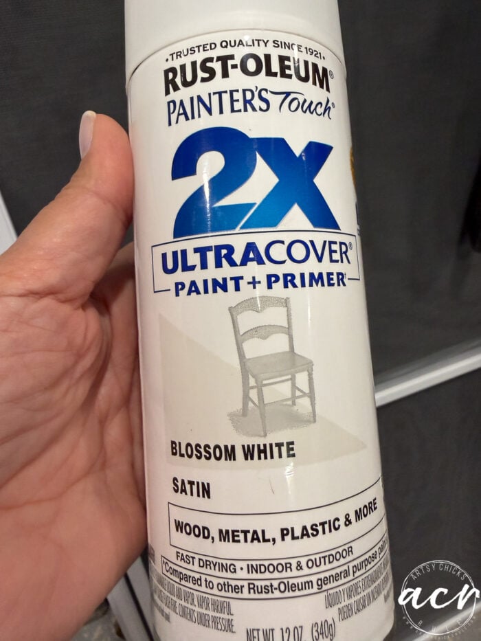 A hand holds a can of Rust-Oleum 2X Ultra Cover paint and primer in blossom white satin finish, designed for wood, metal, plastic, and more—perfect for projects like furniture restoration or giving a petite table a fresh table makeover. A chair graphic is printed on the can.