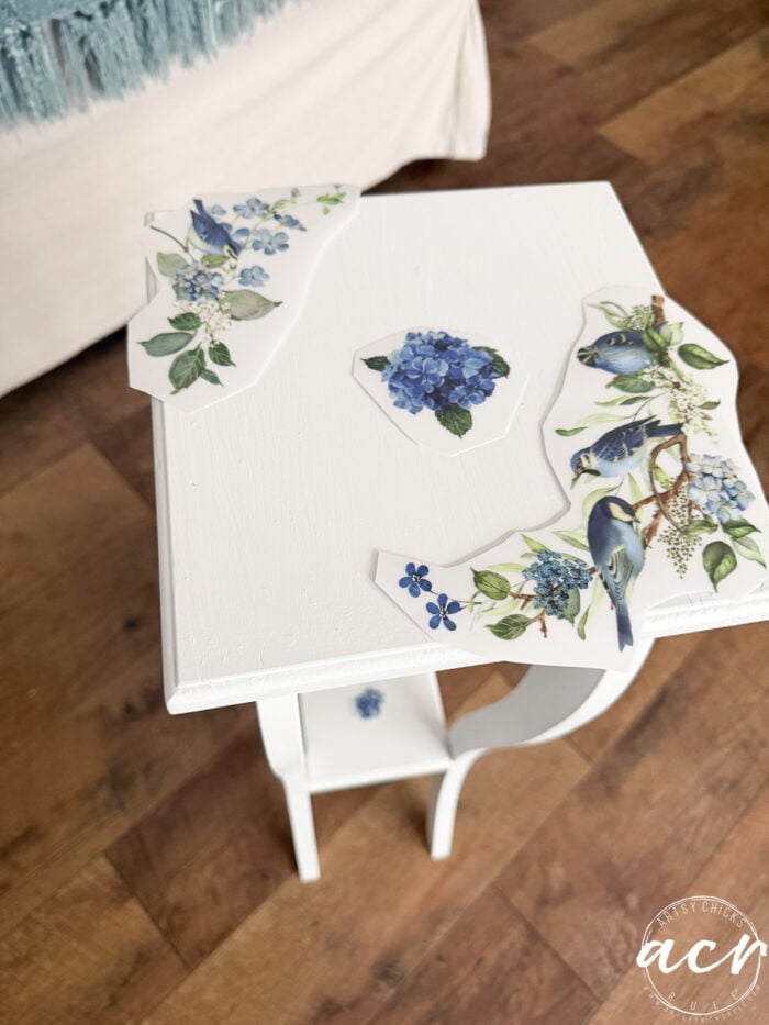 A petite white table with cut-out floral and bird decals arranged on top, ready for application—a perfect setup for a charming table makeover. The floor is wooden, and a white sofa with a blue throw is visible in the background.