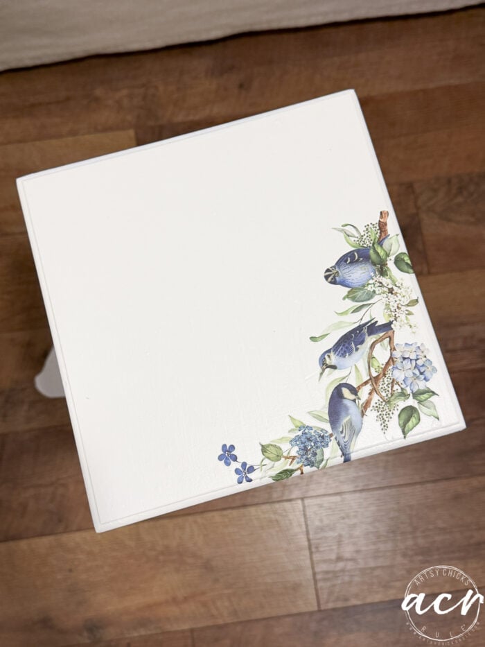 A white square petite table with a floral and bluebird design in one corner, placed on a wooden floor. This charming table makeover features the Artsy Chicks Rule logo on the bottom right corner, highlighting its unique furniture restoration.