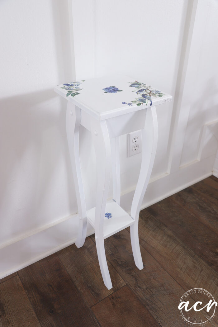 White wooden side table with curved legs, featuring blue floral designs painted on the tabletop, front edge, and lower shelf. Perfect for a furniture makeover, this petite table stands on a dark wood floor against a white paneled wall with an electrical outlet.