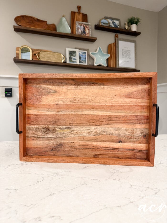 A wooden tray with black handles sits on a marble countertop, perfectly complementing the results of a recent table makeover. In the background, there are floating shelves with decor items, including frames, a star, and plants—a charming display that highlights subtle touches of furniture restoration throughout the space.