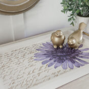 Purple and Gold Tray Makeover A decorative tray with vintage script, a large purple flower, two gold bird figurines, and a small potted plant beside it creates a charming table makeover as it sits on a light wood surface beneath a round mirror—perfect for adding elegance to any petite table.