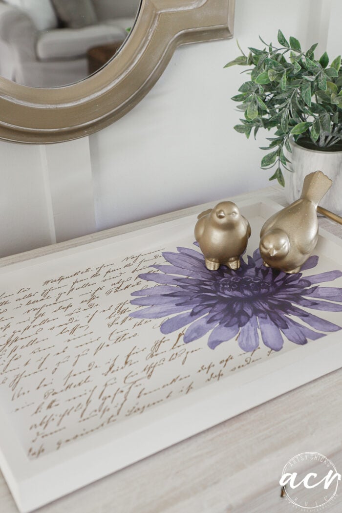 A decorative tray with vintage script, a large purple flower, two gold bird figurines, and a small potted plant beside it creates a charming table makeover as it sits on a light wood surface beneath a round mirror—perfect for adding elegance to any petite table.