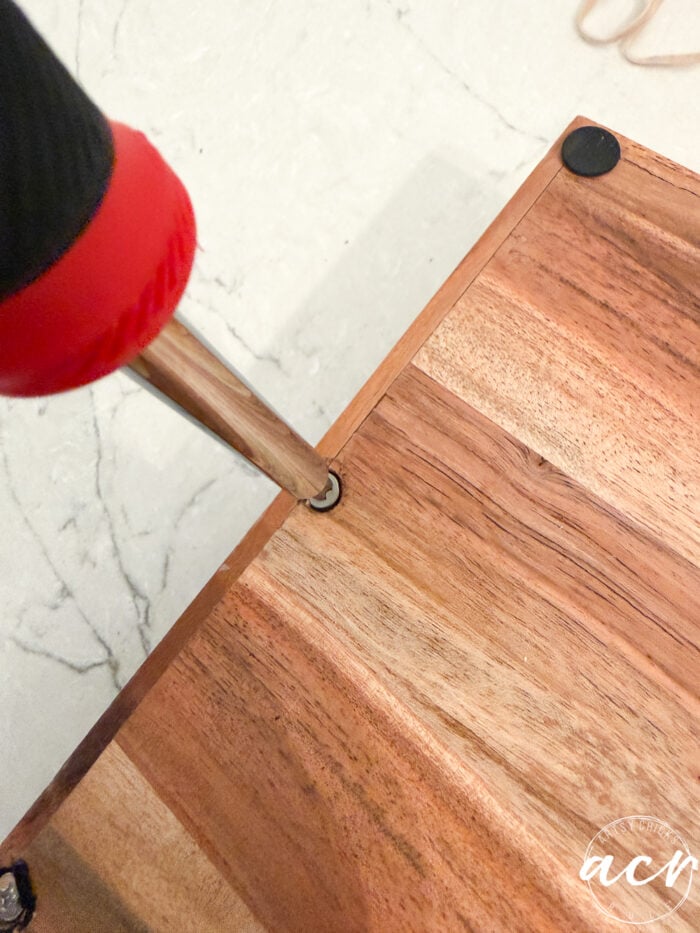 A close-up of a person using a screwdriver to tighten a screw into the corner of a wooden surface on a light marble background, capturing the careful craftsmanship involved in this petite table furniture makeover.