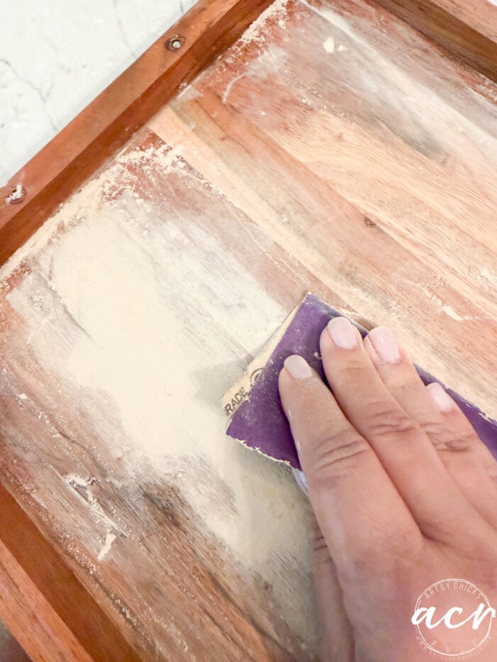 A hand holding sandpaper smooths a wooden surface with visible filler and dust, preparing it for a stunning furniture makeover—perfect for transforming a petite table or beginning an inspired table makeover.