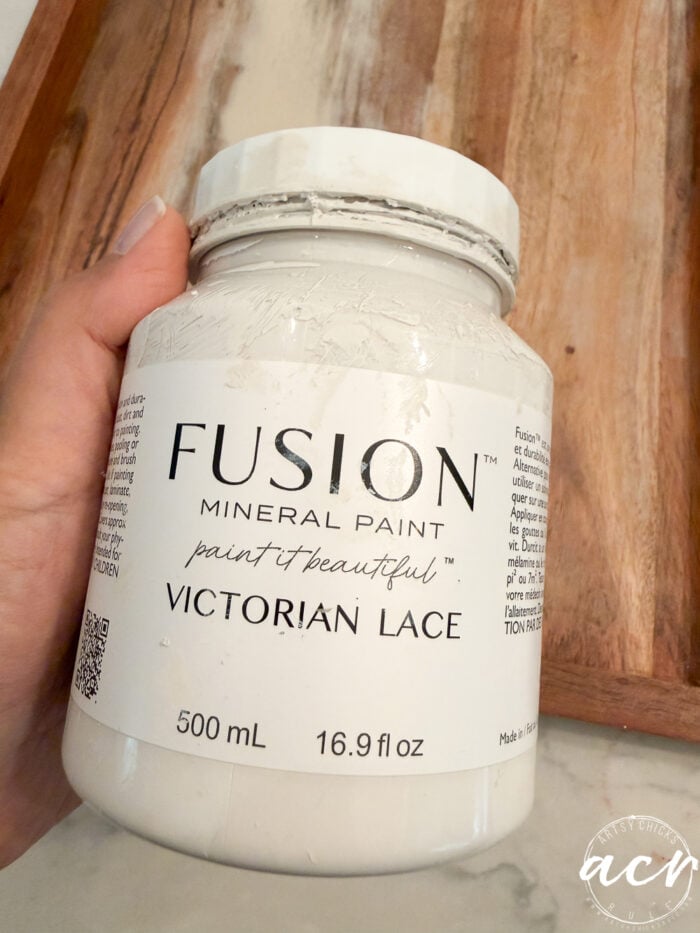 A hand holds a jar of Fusion Mineral Paint in the shade “Victorian Lace,” 500 mL size, perfect for your next petite table or furniture makeover, with a wooden tray in the background.