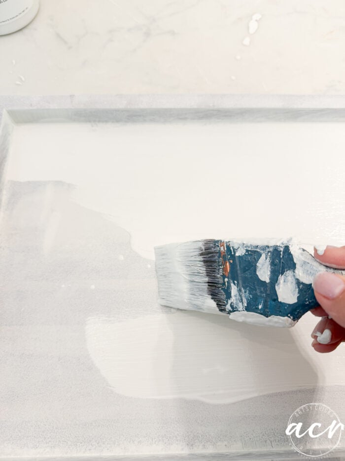 A hand holds a paintbrush applying white paint to a flat wooden surface, capturing the transformative process of a Table Makeover; some paint splatters are visible on the brush and surrounding area, highlighting this moment of Furniture Restoration.