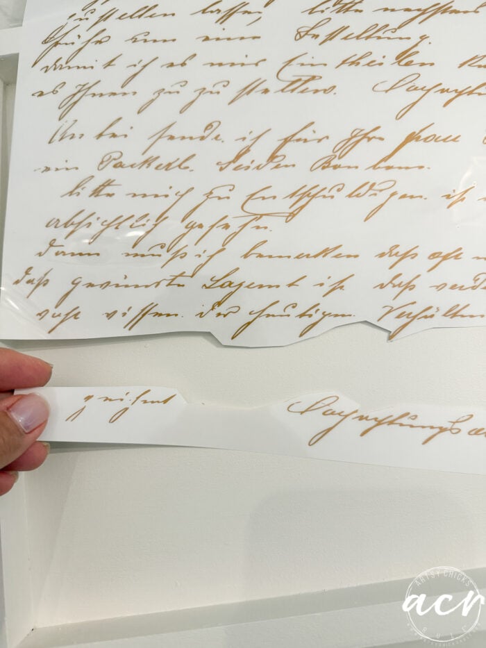 A hand holds a strip of white paper with gold cursive writing, matching a larger sheet with similar script above—perhaps inspiration for an elegant furniture makeover. The text is decorative and partially obscured, suggesting the refined finishing touches often used in a petite table or table makeover project. The bottom right corner has a circular logo with acv.