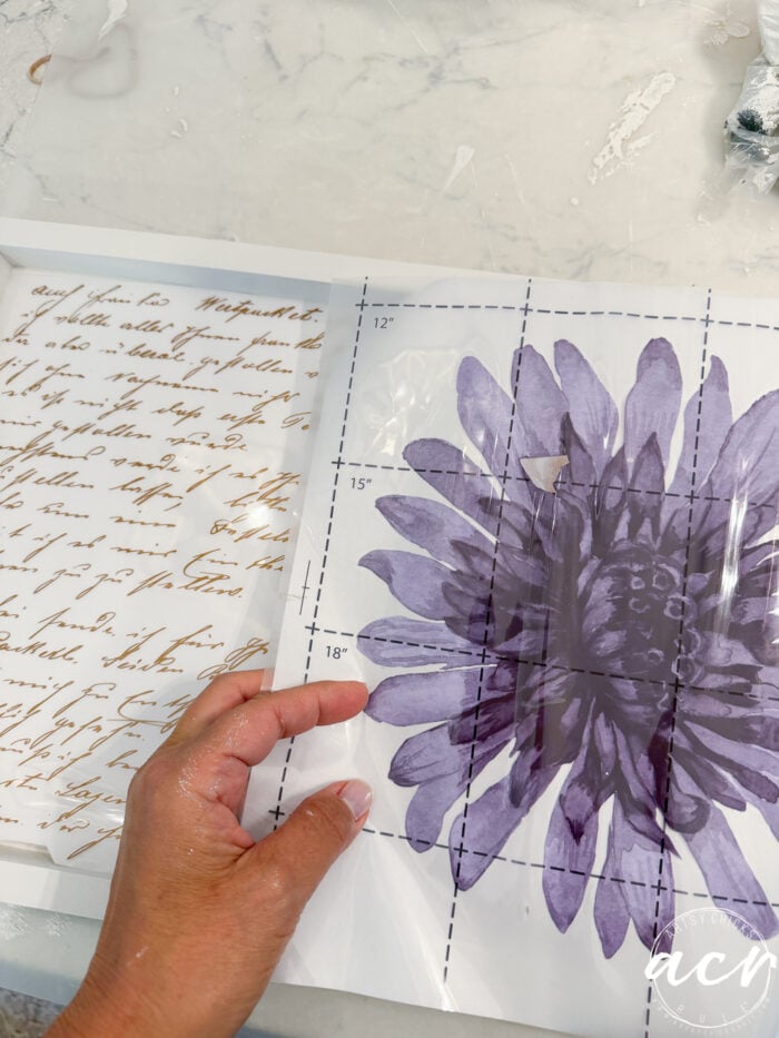 A hand holds a transparent sheet with a large purple flower design over a printed page with cursive writing on a white, marbled surface, hinting at creative ideas for furniture restoration or an upcoming petite table makeover.