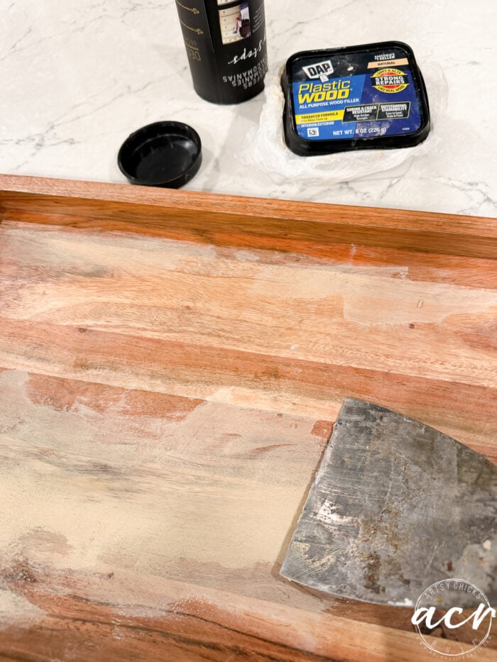 A metal scraper spreads wood filler on a wooden tray during a furniture redo. Nearby are a black coffee cup with a lid and a container of DAP Plastic Wood Filler on a white marble countertop, setting the stage for an inspiring table makeover.