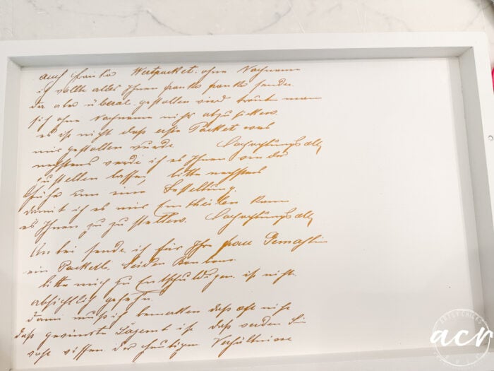 A white-framed piece of art displays elegant brown handwritten script in another language on white paper, set on a marble surface—perfect for adding a touch of sophistication to your next petite table or furniture makeover. The lower right corner has a small circular logo with the letters acy inside.