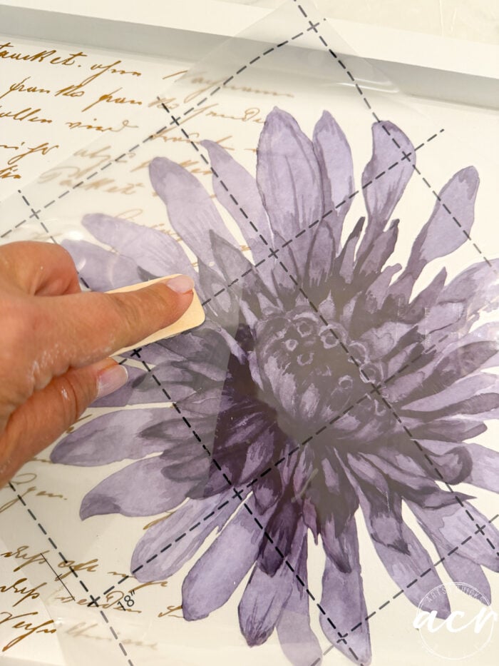 A hand rubs a transfer sheet with a purple flower and script onto a white surface, smoothing it down with a tool during a charming petite table makeover.