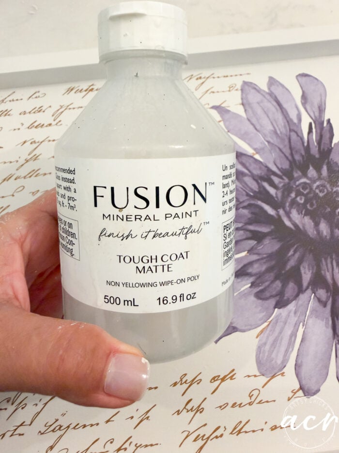A hand holds a 500 mL bottle of Fusion Mineral Paint Tough Coat Matte in front of a floral print with cursive writing in the background, the perfect choice for your next Petite Table or Furniture Makeover.