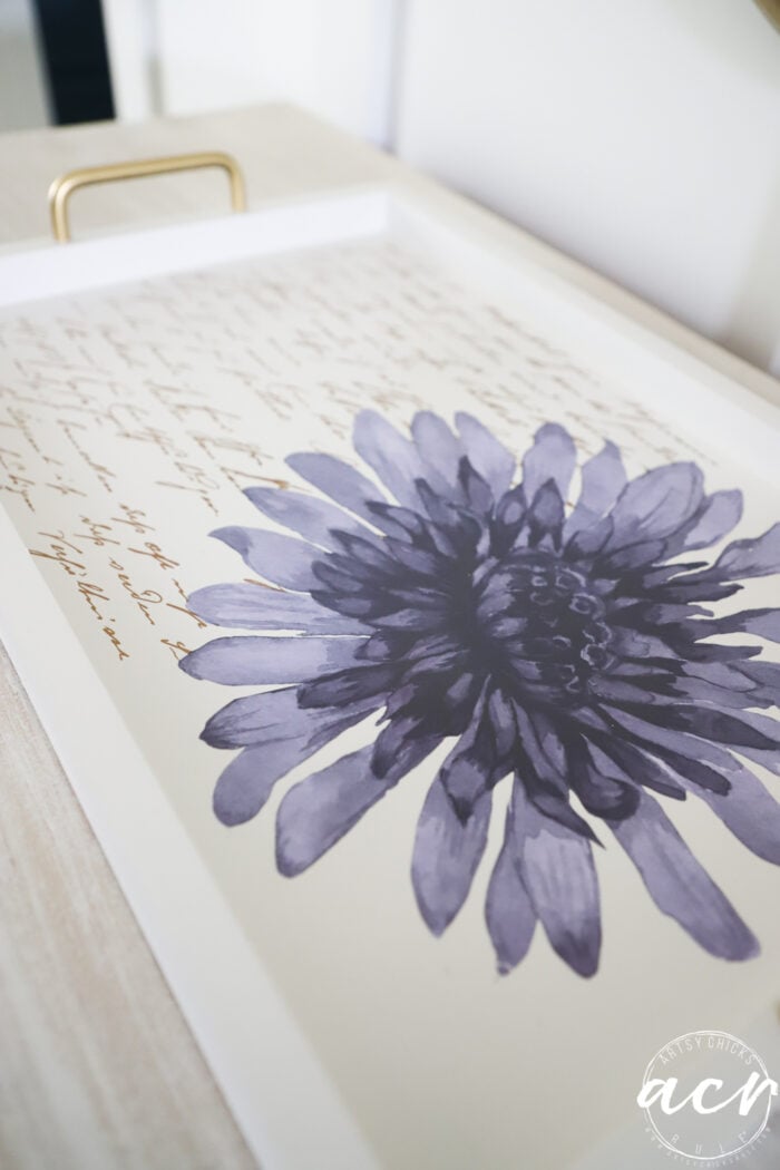 A white tray with gold handles, perfect for a petite table or as part of a furniture makeover, features a large purple flower and script text in the background. The tray rests on a light wooden surface.
