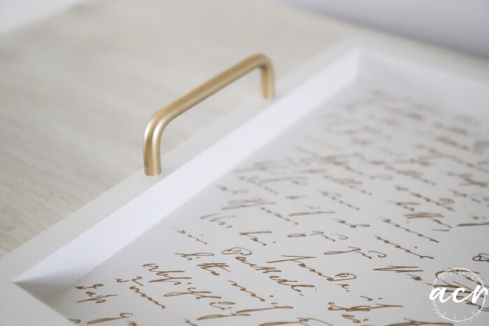 A white tray with a gold handle sits on a light wooden surface, adding a touch of elegance to your Petite Table Makeover. The tray’s bottom features cursive script text in gold, and the lower right corner has a circular logo with the letters acn.
