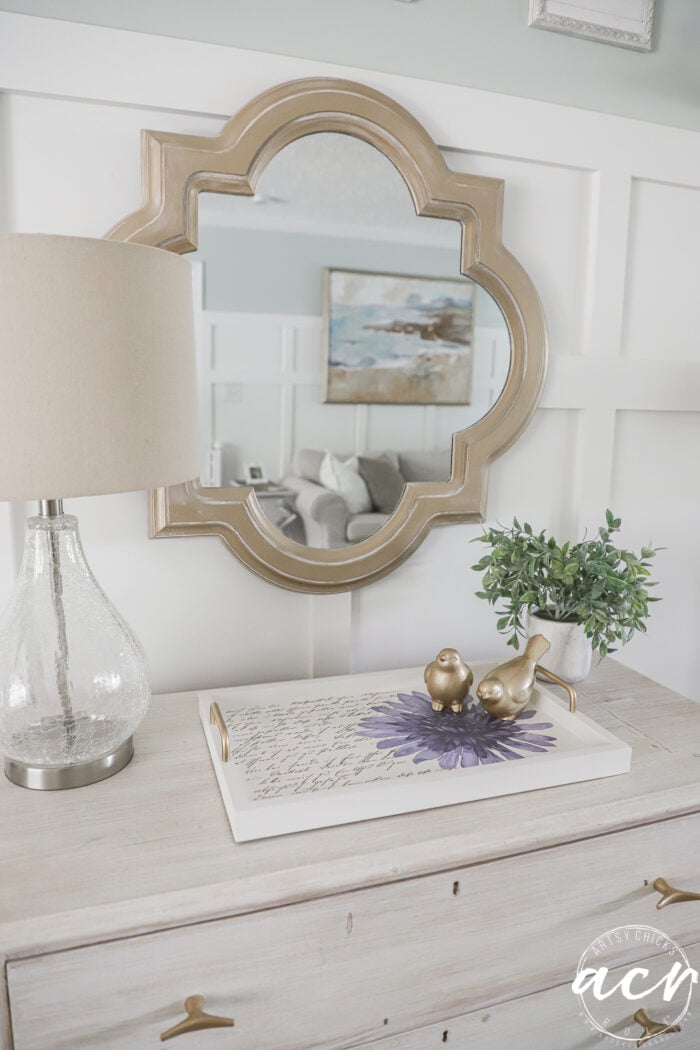 A gold-framed mirror hangs above a light wood dresser that showcases the results of a recent Furniture Makeover, complete with a glass lamp, a leafy plant, and a decorative tray holding gold bird figurines and a purple flower; a living room is reflected in the mirror.