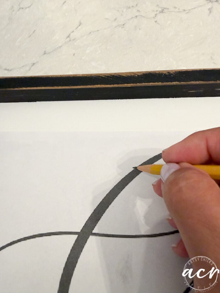 A hand traces a black curved line on white paper with a yellow pencil; a wooden frame—perhaps from a recent thrifted redo—and marble surface are partially visible above.