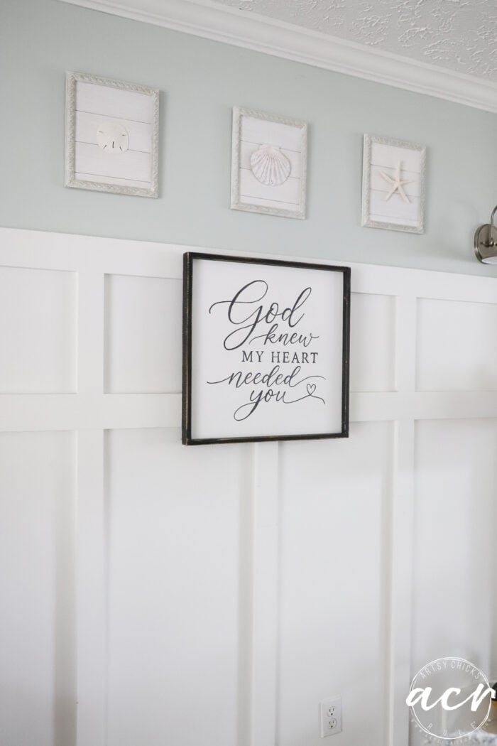 A white wall with four framed artworks: three featuring beach items and one central frame with a thrifted redo of a God Knew My Heart Needed You sign in elegant script.